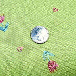 May include: A light green fabric with a textured basketweave pattern featuring small embroidered pink tulips with blue leaves and tiny red hearts. A United States quarter coin is placed in the center for scale, measuring 0.955 inches in diameter.