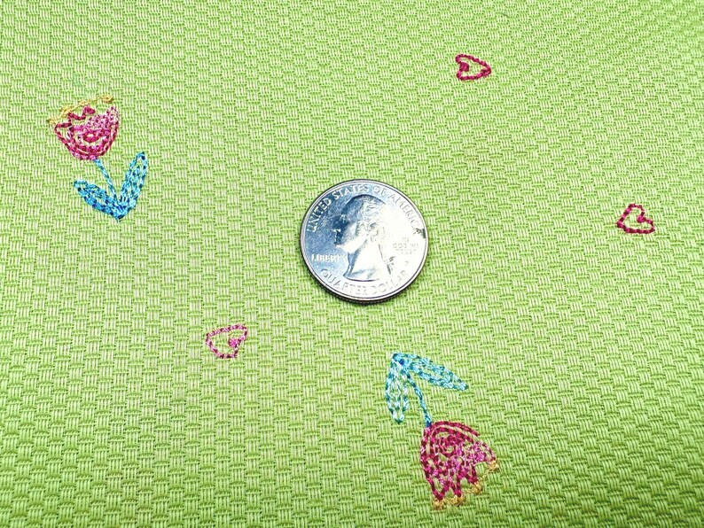 May include: A light green fabric with a textured basketweave pattern featuring small embroidered pink tulips with blue leaves and tiny red hearts. A United States quarter coin is placed in the center for scale, measuring 0.955 inches in diameter.