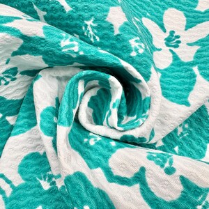 May include: Close-up of a folded fabric with a white background and large teal floral patterns. The fabric has a textured surface and is tightly folded, creating a layered effect. The teal flowers are in full bloom.