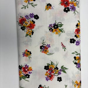 May include: Folded white fabric with a floral pattern. The pattern features clusters of red, yellow, purple, and black flowers with green leaves. The fabric has a textured surface.