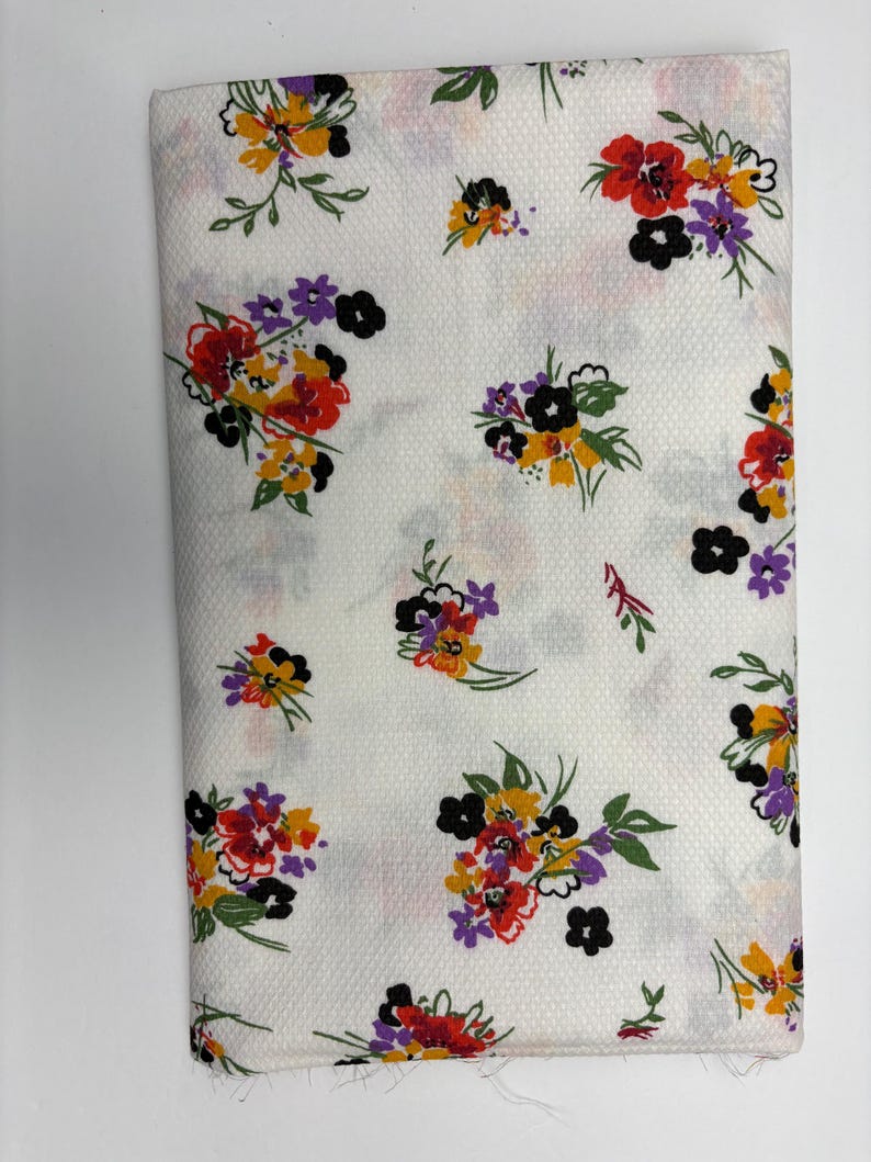 May include: Folded white fabric with a floral pattern. The pattern features clusters of red, yellow, purple, and black flowers with green leaves. The fabric has a textured surface.