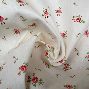 Puede incluir: Tela blanquecina con un patrón repetido de pequeñas rosas rosas y hojas verdes. La tela está ligeramente arrugada, creando sombras suaves y profundidad. El diseño floral está distribuido uniformemente.