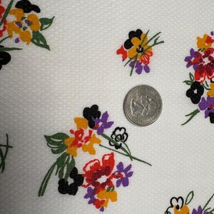 May include: White fabric with a repeating floral pattern. The flowers are in clusters of red, yellow, purple, and black. A US quarter is included for scale, showing the fabric's texture and the size of the floral elements.