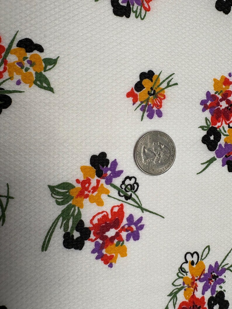 May include: White fabric with a repeating floral pattern. The flowers are in clusters of red, yellow, purple, and black. A US quarter is included for scale, showing the fabric's texture and the size of the floral elements.