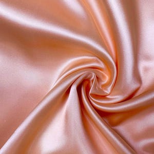 May include: Close-up of a peach-colored satin fabric with soft folds and a lustrous sheen. The fabric is tightly woven, creating a smooth surface. The light reflects off the fabric, highlighting the curves and textures of the folds.