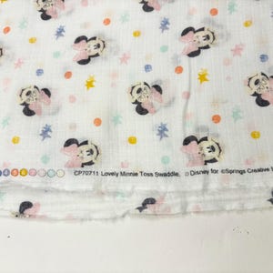 Op de afbeelding: Witte stof met een herhaald patroon van Minnie Mouse, roze strikken en kleurrijke stippen en sterren. De tekst "Lovely Minnie Toss Swaddle" en "Disney" is op de stof gedrukt.