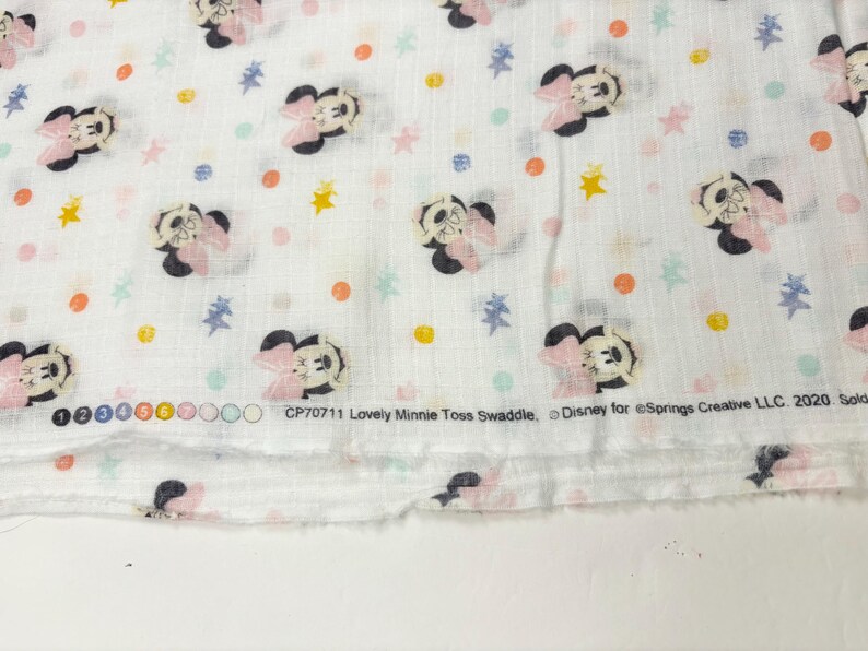 Op de afbeelding: Witte stof met een herhaald patroon van Minnie Mouse, roze strikken en kleurrijke stippen en sterren. De tekst "Lovely Minnie Toss Swaddle" en "Disney" is op de stof gedrukt.