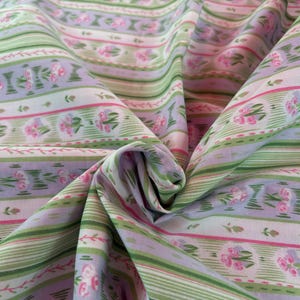 Könnte beinhalten: Gefalteter Stoff mit einem sich wiederholenden Muster aus grünen und weißen Streifen, unterbrochen von Lavendelbändern und rosa Blumensträußen. Die Textur und der Fall des Stoffes sind sichtbar. Die Farbpalette ist weich und pastellfarben.
