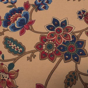 Op de afbeelding: Close-up van een beige stof met een bloemenpatroon. Het ontwerp toont gestileerde bloemen in tinten blauw, rood en bruin, met groene accenten. De bloemen zijn verbonden door bruine stelen, waardoor een herhalend patroon ontstaat.