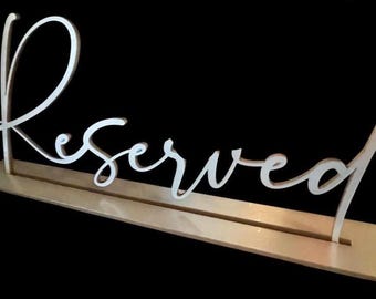 May include: A light-colored wooden sign featuring the word Reserved written in a white cursive script font. The sign consists of a flat rectangular base with a slot that holds the upright cutout lettering securely in place.