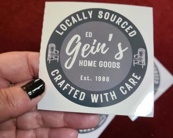 May include: A white square sticker with a circular design featuring the text "Locally Sourced" and "Crafted With Care". The center circle has "Ed Gein's Home Goods" and "Est. 1906". Two chair illustrations flank the text.