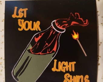 May include: A black and green glass bottle with a red and orange flame coming out of the top. The bottle is labeled "Let Your Light Shine" in orange text.