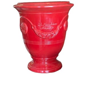 May include: A vibrant red ceramic planter with a glossy finish. The planter features embossed decorative elements and the text "La Madeleine Vintage". It has a wide top, a rounded body, and a pedestal base, suitable for indoor or outdoor use.