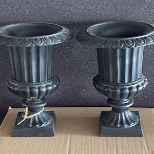 May include: Two black, decorative urns with fluted bodies and ornate detailing. Each urn has a wide, scalloped rim and sits on a square base. A natural twine is tied around the base of one urn. These urns are suitable for indoor or outdoor use.