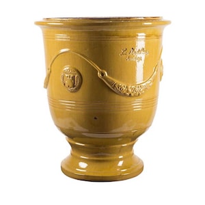 May include: Mustard yellow ceramic planter with a pedestal base. The planter features embossed decorative elements, including a crest and floral designs. The text "La Madura Andiga" is visible on the side. A classic piece for home decor.