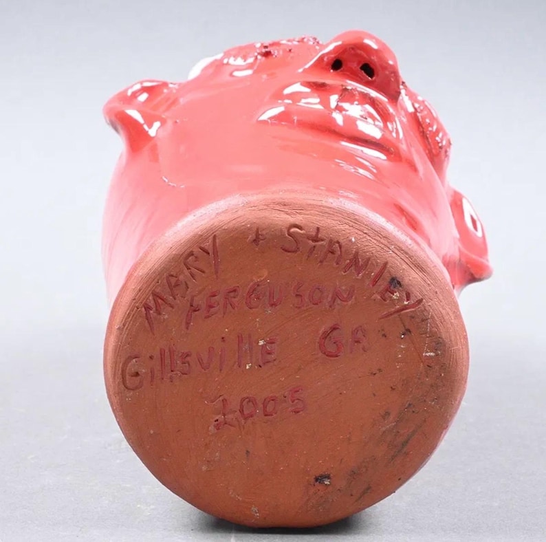 Puede incluir: Una taza de cer&aacute;mica roja con forma de rostro con nariz, boca y ojos detallados. La base de la taza tiene texto escrito a mano que dice "MARY + STANLEY FERGUSON Gillsville GA 2005".