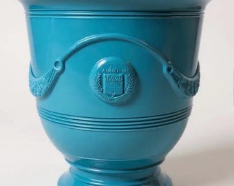 May include: A glossy turquoise ceramic planter with a wide flared rim and a pedestal base. The exterior features horizontal ribbed detailing and a central circular medallion embossed with a book icon surrounded by laurel leaves and draped swags.