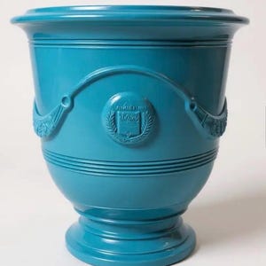 May include: A glossy turquoise ceramic planter with a wide flared rim and a pedestal base. The exterior features horizontal ribbed detailing and a central circular medallion embossed with a book icon surrounded by laurel leaves and draped swags.