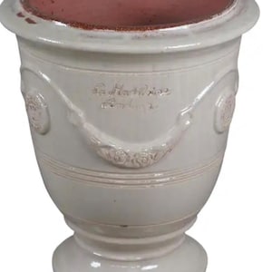 May include: A cream-colored ceramic planter with a wide, flared rim and a pedestal base. The exterior features embossed floral swags and circular medallions. The interior is a contrasting terracotta red. The text La Madeleine is embossed on the front.