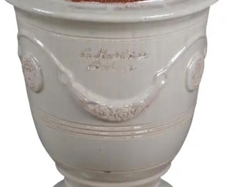 May include: A cream-colored ceramic planter with a wide, flared rim and a pedestal base. The exterior features embossed floral swags and circular medallions. The interior is a contrasting terracotta red. The text La Madeleine is embossed on the front.