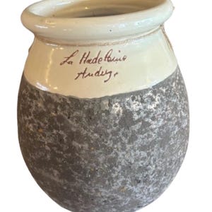 May include: A small, handcrafted ceramic pot with a textured, speckled gray lower half and a cream-colored upper section. The pot has a slightly flared rim and features the handwritten text "La Hadelaine Audey".