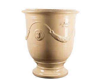May include: A cream-colored ceramic planter with a flared rim and a pedestal base. The exterior features a raised circular emblem depicting a castle and a decorative swag motif with floral details, along with the embossed text La Madeleine.