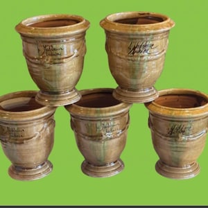 May include: Five brown ceramic planters with a textured finish and a wide rim. Each planter has a cylindrical shape with a decorative band and a pedestal base. The planters have a handwritten inscription. The background is green.