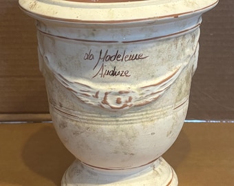 May include: A cream-colored terracotta vase with a weathered finish, featuring a flared rim, a pedestal base, and a decorative garland relief. The front is inscribed with the handwritten text 'la Madeleine Anduze' in a dark brown script.