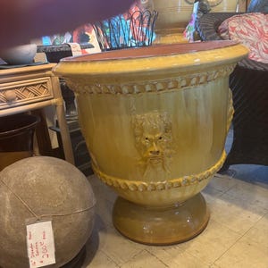 May include: A large, yellow-glazed ceramic planter with a detailed face design. The planter has a wide rim and a pedestal base. A stone sphere is visible in the foreground. Other planters are in the background.