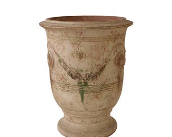 May include: A large, weathered cream-colored urn with a wide, flared rim and a pedestal base. The exterior features a distressed finish with patches of green and brown, decorated with embossed floral swags and circular medallions around the body.