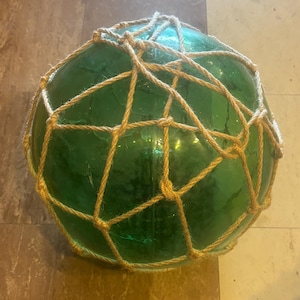 May include: A large, spherical, translucent green glass fishing float encased in a decorative, hand-tied tan jute rope netting. The glass surface is smooth with visible internal bubbles and a distinct vertical seam running across the center.