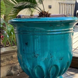 Puede incluir: Una gran maceta de cerámica turquesa con un acabado envejecido. La maceta tiene una parte superior ancha y acanalada y una base decorativa. Una pequeña palmera está plantada dentro de la maceta. La maceta está al aire libre en una terraza de madera.