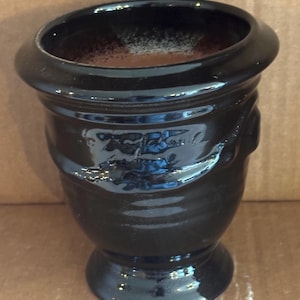 May include: A glossy black ceramic mate cup with a wide, rounded rim and a pedestal base. The exterior features a subtle, embossed decorative band around the upper section, and the interior is filled with a dark brown, textured substance.