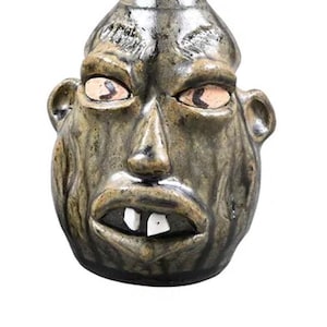 May include: A ceramic face jug with a grotesque expression. The jug is glazed in shades of brown and green, with prominent features including bulging eyes, a large nose, ears, and two prominent teeth. The jug has a small opening at the top.