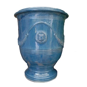 May include: A glossy blue ceramic planter with a wide flared rim and a tapered base. The exterior features a raised circular emblem with text and decorative swags draped across the upper body, showcasing a smooth, reflective glazed finish.