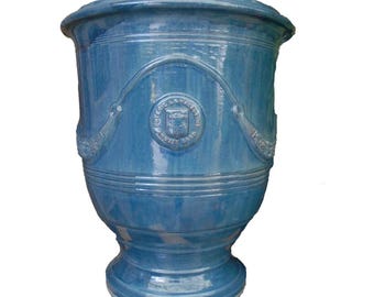 May include: A glossy blue ceramic planter with a wide flared rim and a tapered base. The exterior features a raised circular emblem with text and decorative swags draped across the upper body, showcasing a smooth, reflective glazed finish.