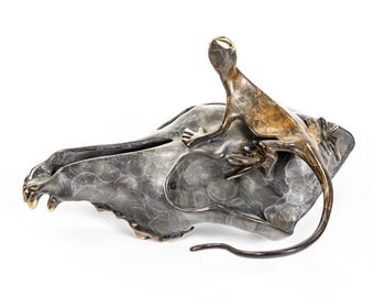 May include: A bronze sculpture of a lizard perched on a skull-shaped bowl. The lizard has a detailed texture and a long tail. The bowl has a textured, mottled surface. The sculpture is set against a white background.