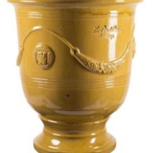 May include: A mustard yellow glazed ceramic planter with a wide rim and a pedestal base. The exterior features embossed decorative swags and circular medallions. The surface has a glossy finish with subtle text markings visible on the upper body.