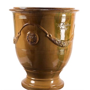 May include: A glazed ceramic planter in a warm ochre brown hue with a glossy finish. The vessel features a flared rim, a pedestal base, and embossed decorative swags and a circular crest. The text La Madeleine Anduze is visible on the side.