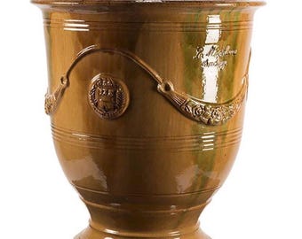 May include: A glazed ceramic planter in a warm ochre brown hue with a glossy finish. The vessel features a flared rim, a pedestal base, and embossed decorative swags and a circular crest. The text La Madeleine Anduze is visible on the side.