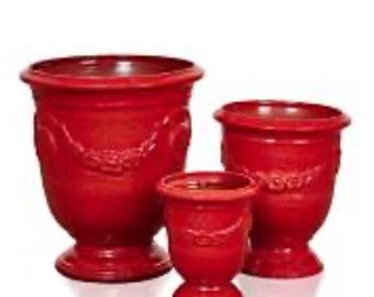 May include: A set of three glossy red planters of varying sizes, each featuring a wide rim, a pedestal base, and a decorative embossed garland pattern encircling the upper body of the vessel. The planters are arranged in a cluster against a white background.