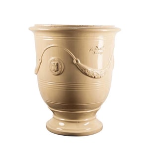 May include: A cream-colored ceramic planter with a flared rim and a pedestal base. The exterior features a raised circular emblem depicting a castle and a decorative swag motif with floral details, along with the embossed text La Madeleine.