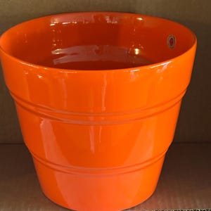May include: A bright orange ceramic plant pot with a glossy finish. The pot features a wide, open circular top and a tapered body with two horizontal, recessed ring details encircling the exterior. A small circular sticker is visible inside.