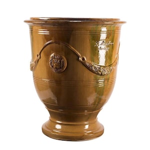 May include: A glazed brown ceramic planter with a flared rim and a pedestal base. The exterior features a raised relief design of draped garlands and a circular crest. The text "La Madeleine Anduze" is embossed on the upper side of the vessel.