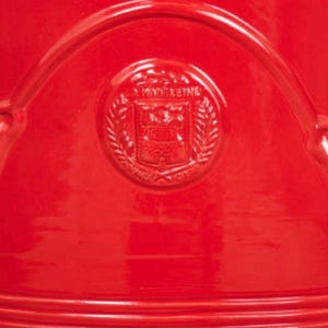 May include: A bright red ceramic surface featuring a circular embossed emblem in the center. The emblem displays the text LA MADELEINE surrounded by a laurel wreath design. The object has a smooth, glossy finish with curved, raised lines.