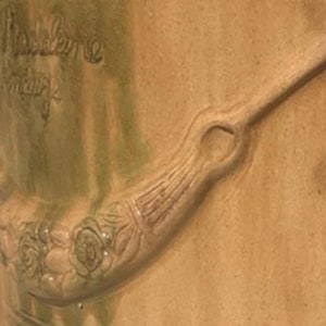 May include: Close-up of a ceramic vase with a handle and floral detailing. The handle features a series of carved lines and a circular opening. The vase has a light brown color with green accents and the text "Madeleine" and "Hendrik" is visible.
