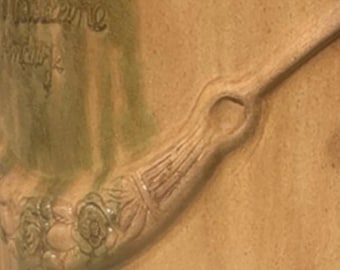 May include: Close-up of a ceramic vase with a handle and floral detailing. The handle features a series of carved lines and a circular opening. The vase has a light brown color with green accents and the text "Madeleine" and "Hendrik" is visible.