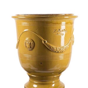 May include: A mustard yellow glazed ceramic planter with a flared rim and a pedestal base. The exterior features embossed swags, floral garlands, a circular crest with a castle motif, and the embossed text La Madeleine Anduze.