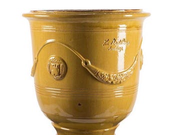 May include: A mustard yellow glazed ceramic planter with a wide rim and a pedestal base. The exterior features embossed swags, floral details, and a circular medallion with a castle emblem. The text La Madeleine Anduze is embossed on the side.