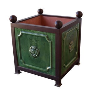 May include: A square, green ceramic planter with a dark brown frame and accents. The planter features embossed details, a crest, and text. Four dark brown spheres adorn the top corners. The interior is a reddish-brown color.